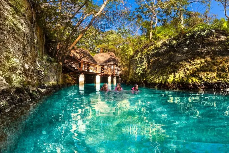 Turquoise natural pool with swimmers beside a wooden pavilion at Xcaret park near Playa del Carmen, Mexico